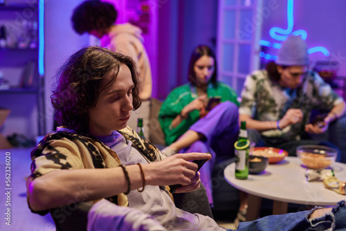 Young restful man in casualwear sitting in armchair against his friends in living room lit by neon light at home party and using mobile phone