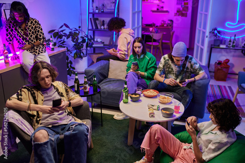 Group of young multicultural friends in casualwear using mobile gadgets while sitting on couch and in armchairs in living room lit by neon light