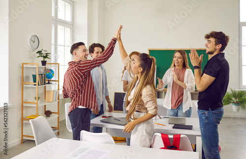 Cheerful college students give each other high five while celebrating success of their project. Two satisfied student friends in classroom congratulate each other to applause of classmates.