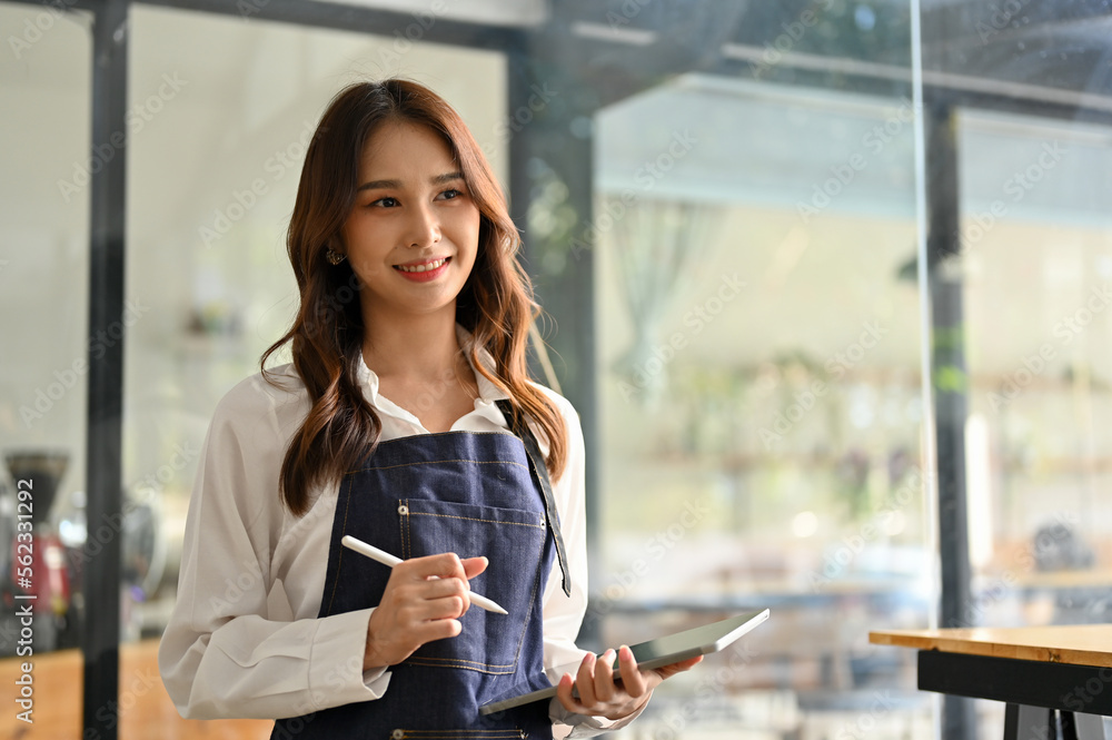 Attractive Asian female waitress or coffee shop staff in apron holding a tablet, standing in her cafe. - obrazy, fototapety, plakaty 