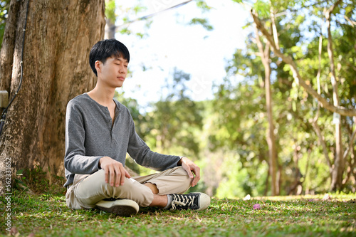 Obraz na plátně Relaxed young Asian man in casual clothes meditating under the tree in the greenery park