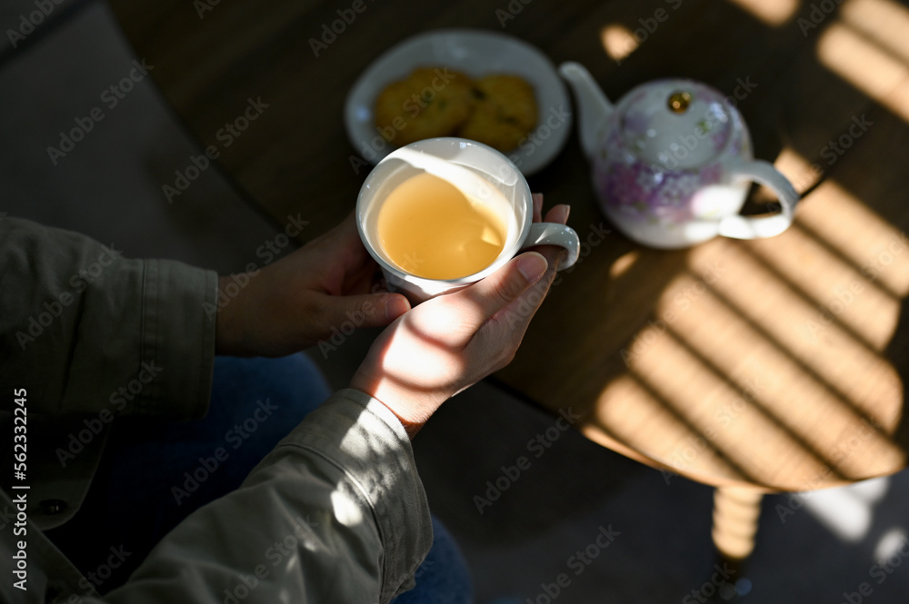 A woman's hands holding a cup of hot tea over blurred tea table in the background.
