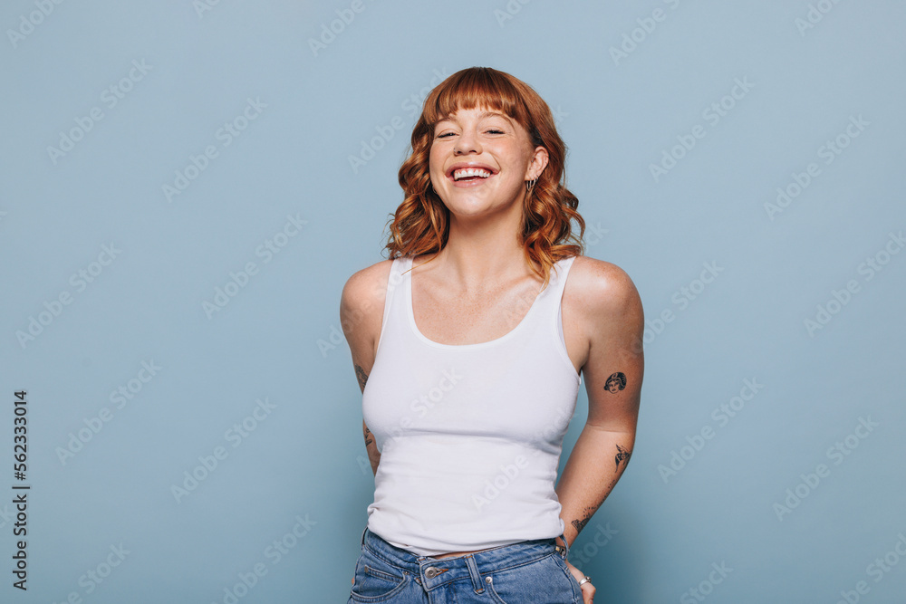 Cheerful young woman smiling at the camera while standing in a tank top and jeans - obrazy, fototapety, plakaty 
