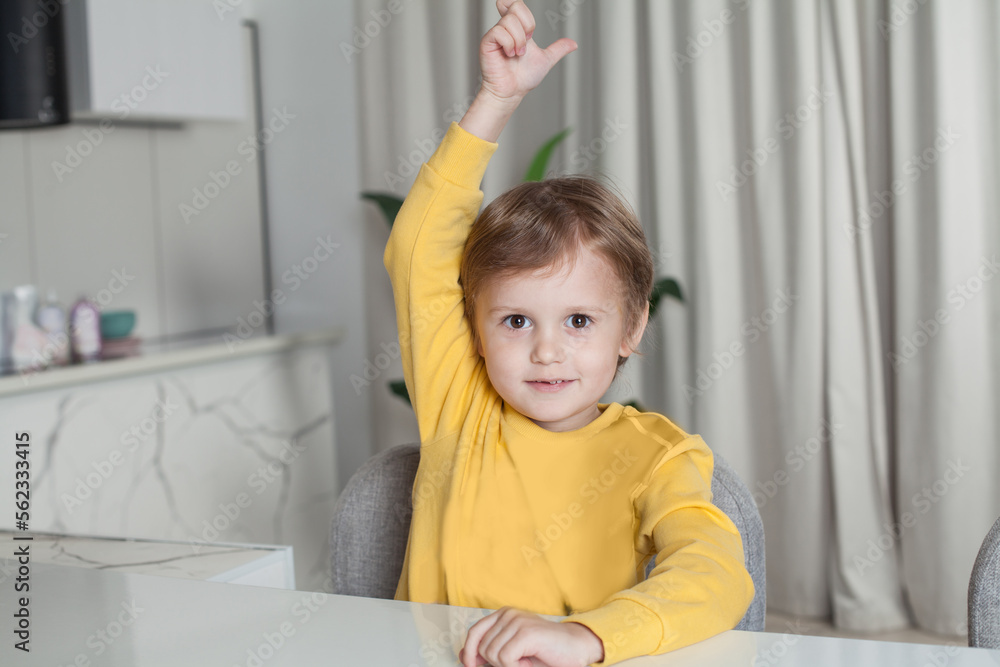 Smiling happy child boy in yellow sweater sitting by the table and raising his hand up with thumb up on studio kitchen background Smiling happy child boy in yellow sweater sitting by the table and raising his hand up with thumb up on studio kitchen background