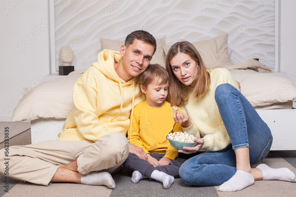 Friendly cute family, parents in yellow clothes with child son sitting by the bed and eating popcorn from blue bowl