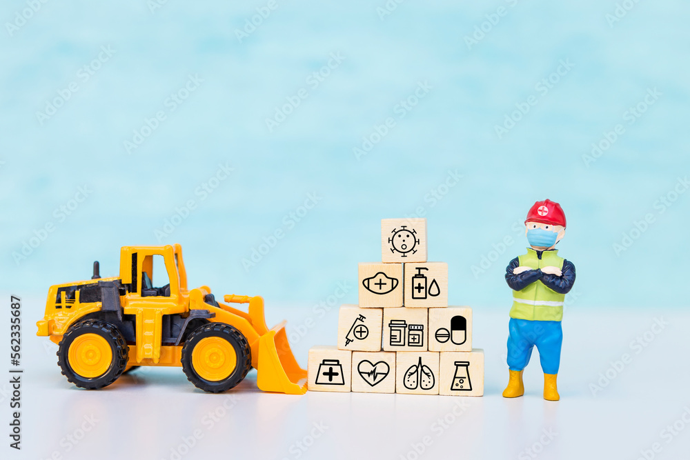 Obraz premium Miniature worker wearing safety helpmets with medical icon cube and yellow front loader truck with space on blue background, health and medical concept
