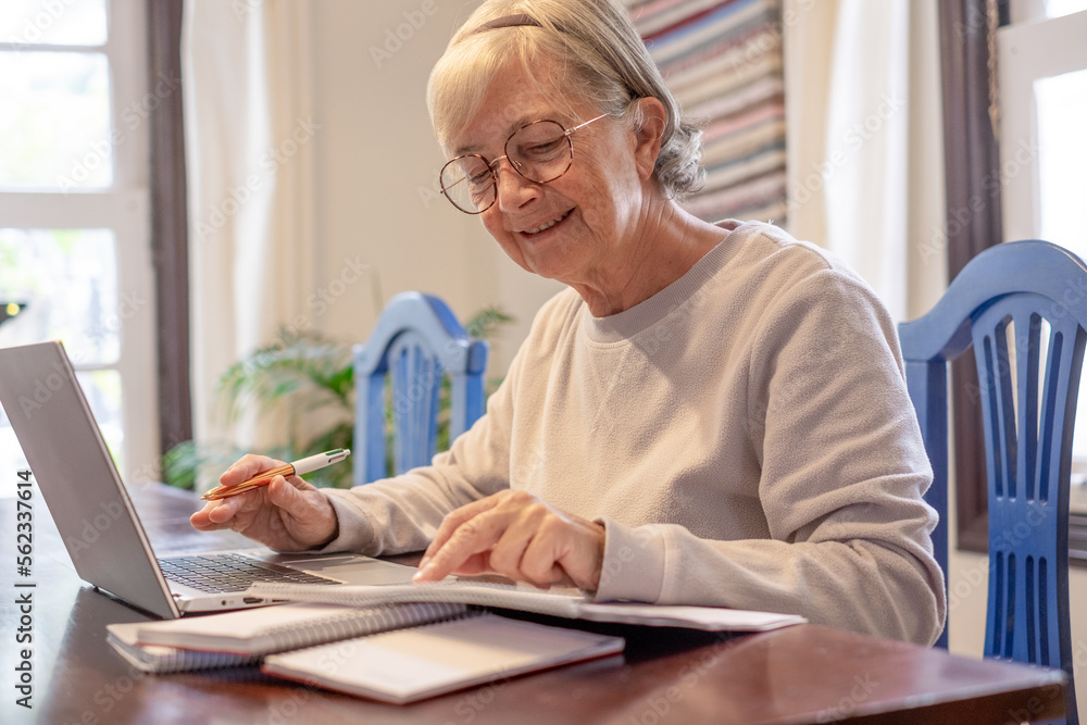 Smiling mature woman sitting at table with laptop and books working or studying. Attractive senior woman in eyeglasses enjoying teaching activity and learning using new technology Smiling mature woman sitting at table with laptop and books working or studying. Attractive senior woman in eyeglasses enjoying teaching activity and learning using new technology