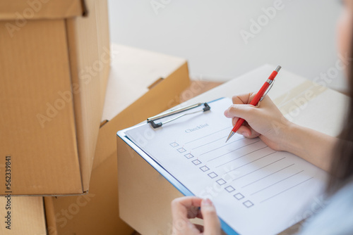 Asian woman checking and packing the carton box prepare to move to new house relocation shipping or check goods package before delivery to customer from online shopping by checklist