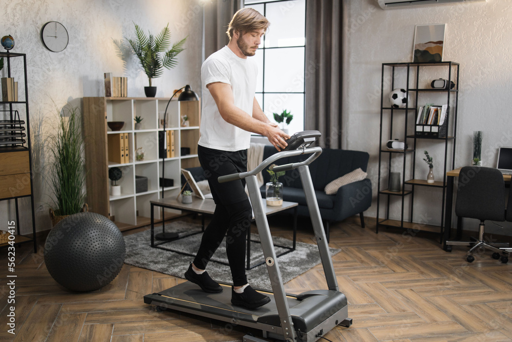 Fototapeta premium Full length view of blond sporty man in white t-shirt and black sports pants having cardio training to loss weight using treadmill at home gym.