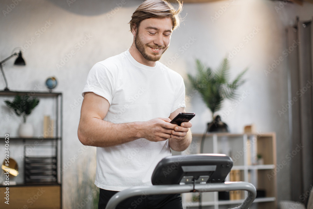 Handsome young sports man using phone looking online fitness class while working out, running, doing cardio training on treadmill in morning time, indoor on background of modern apartment Handsome young sports man using phone looking online fitness class while working out, running, doing cardio training on treadmill in morning time, indoor on background of modern apartment
