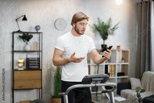 Front view of caucasian man in sports clothes using smart phone for video call doing cardio training on treadmill at home or gym. Concept of sport, health care, action, remote leisure.