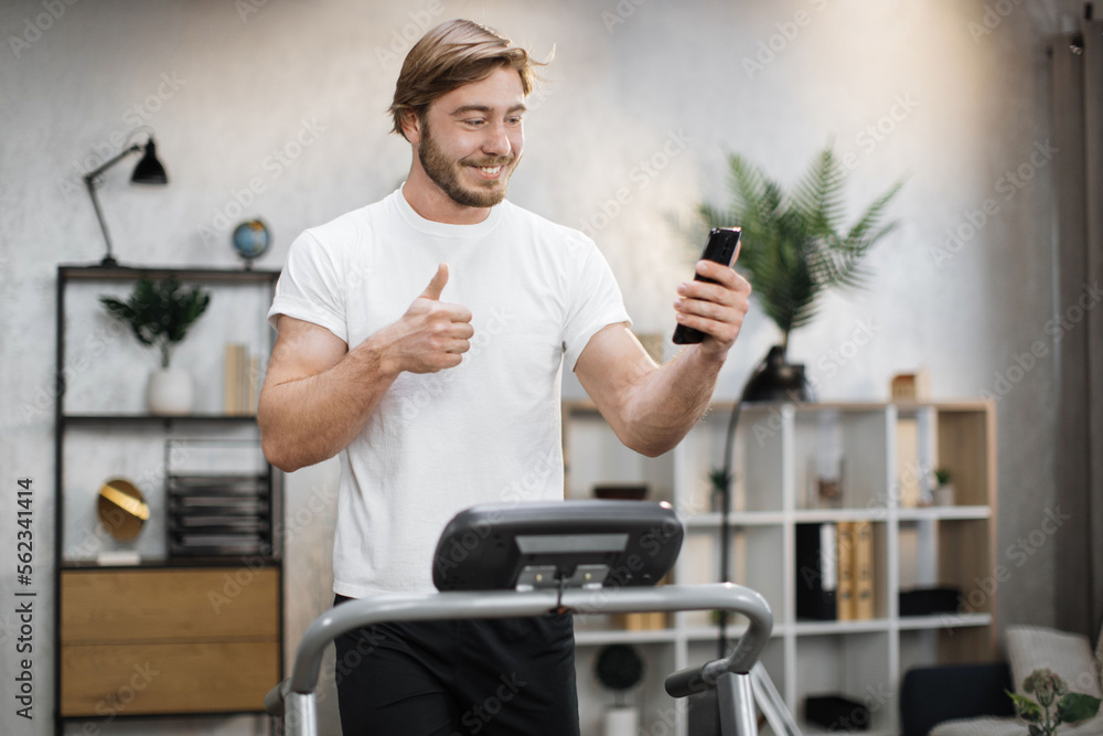 Close up view of bearded young sporty man using running machine simulator while making selfie or having video call showing thumb up. Blond sportsman having workout on treadmill indoors his home.