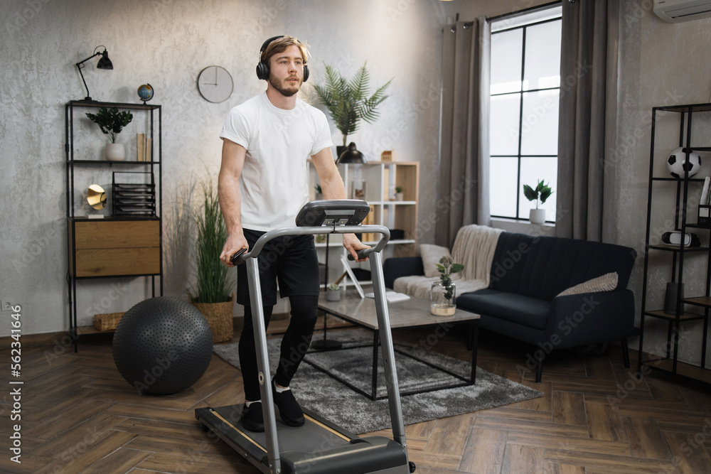 Happy young sporty male athlete jogging on treadmill using phone at home. Handsome muscular, fit man running while listening music in headphones while training during leisure activity.