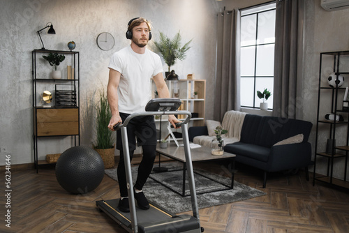 Happy young sporty male athlete jogging on treadmill using phone at home. Handsome muscular, fit man running while listening music in headphones while training during leisure activity. © sofiko14