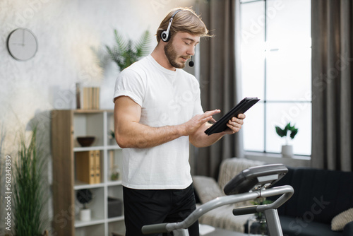 Smiling muscular sporty businessman in headset working remote from home using digital tablet, while having cardio workout, running on treadmill.