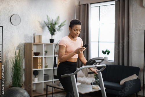 Portrait of active african woman in sportswear using smartphone, running using treadmill at home at morning. Coronavirus Covid 19 social distance. Home workout, stay at home, remote leisure concept.