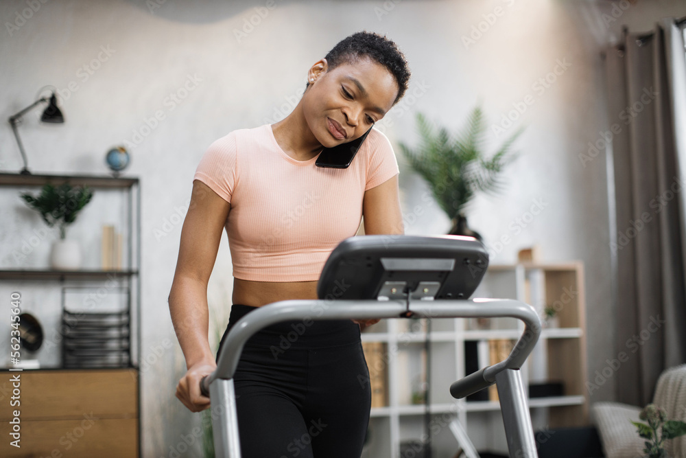 Front view of african sporty woman in pink t-shirt working out in treadmill while having phone call with her relatives or friends. Home activity, lifestyle concept. Front view of african sporty woman in pink t-shirt working out in treadmill while having phone call with her relatives or friends. Home activity, lifestyle concept.
