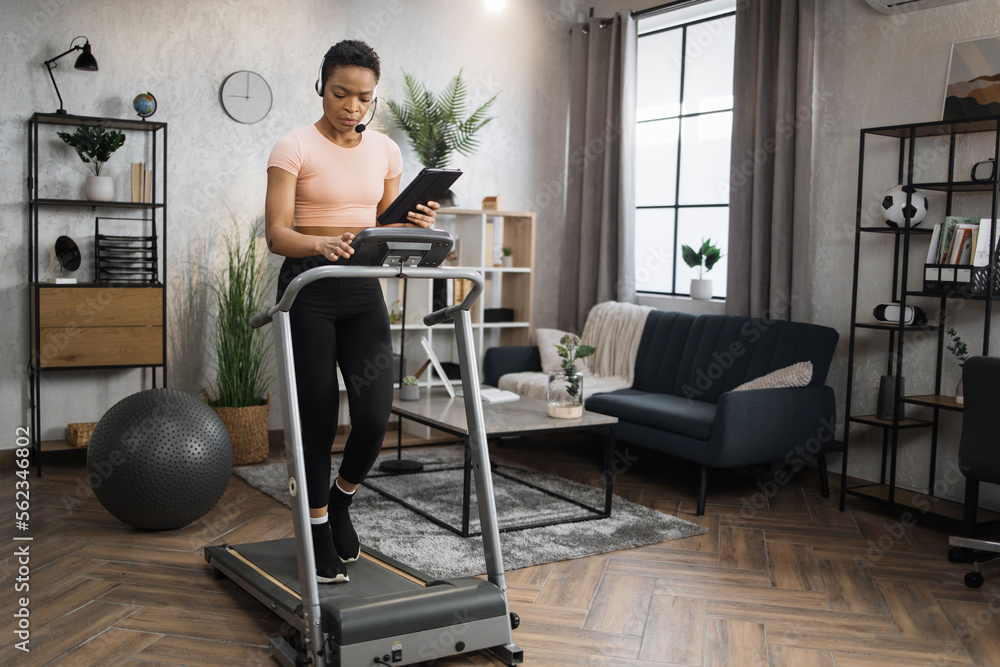 Fototapeta premium Side view of african woman in sport clothes using headset working on tablet computer while doing cardio training on treadmill at morning at home gym. Concept of sport, health care, business.
