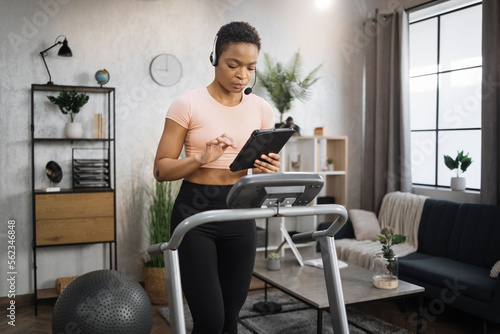 Confident muscular sporty businesswoman in headset working remote from home using digital tablet  while having cardio workout  running on treadmill.