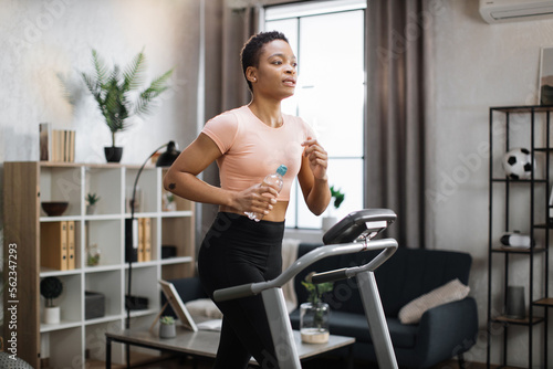 Attractive young sports african american woman holding bottle with water working out, running, doing cardio training on treadmill, indoor on background of modern apartment.