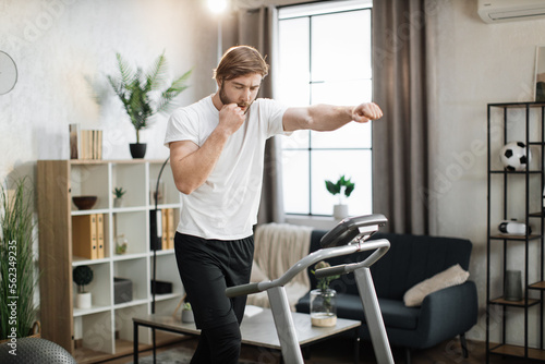 Strong young caucasian man doing boxing punches while training indoors. Professional bearded muscle sportsman boxing and exercising warm-up during home workout at treadmill.