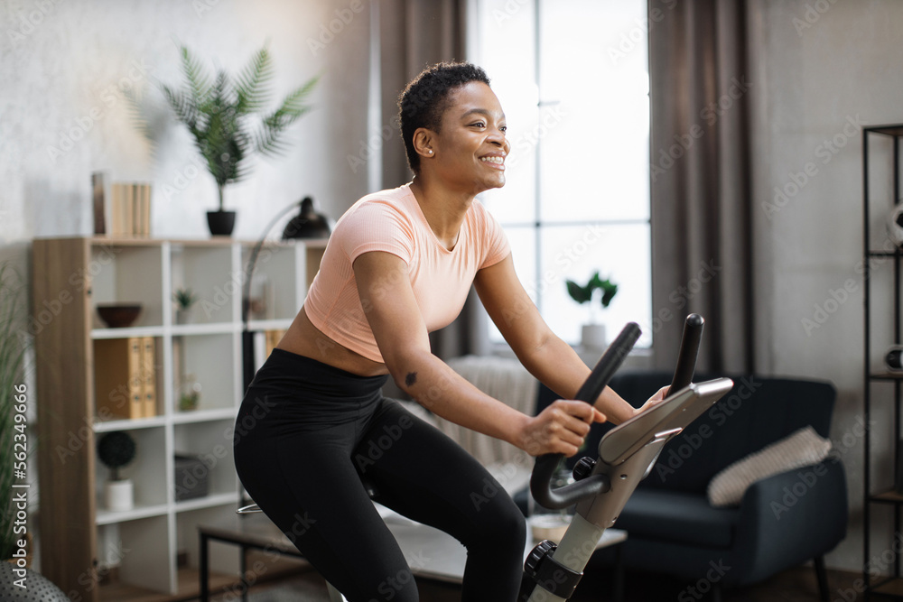 Smiling beautiful african american sports woman in sportswear cycling bike at home on background of light living room. Cardio training, exercising legs, cardio workout indoors. - obrazy, fototapety, plakaty 