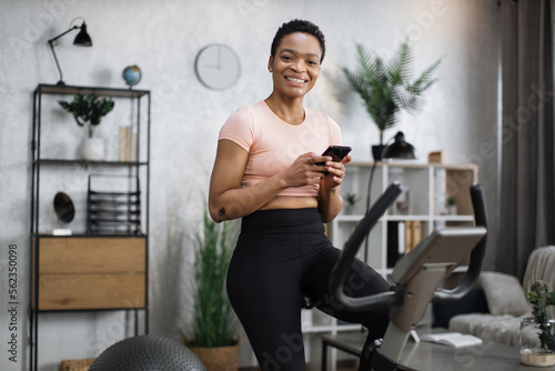 Portrait of young african female wearing sportswear writing message on her smartphone while working out using exercise bike. Home fitness workout sporty woman training on smart stationary bike indoors