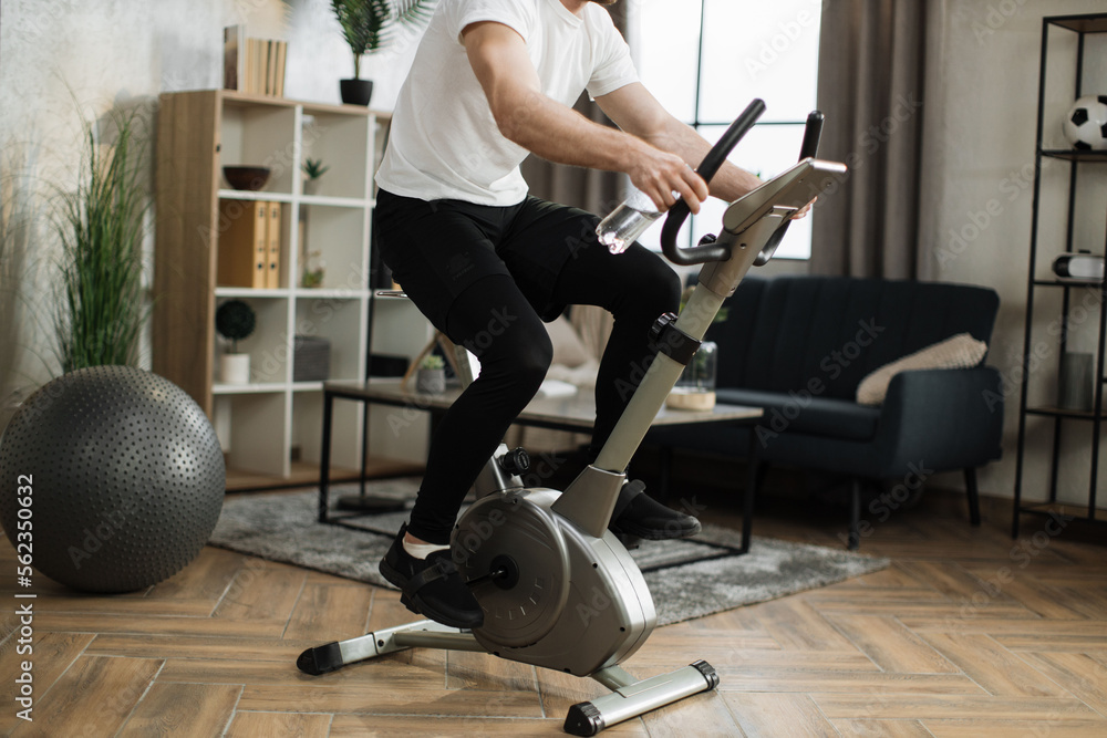 Cropped view of young male with water bottle wearing sportswear using exercise bike. Home fitness workout sporty man training on smart stationary bike indoors. Young caucasian guy athlete. Cropped view of young male with water bottle wearing sportswear using exercise bike. Home fitness workout sporty man training on smart stationary bike indoors. Young caucasian guy athlete.