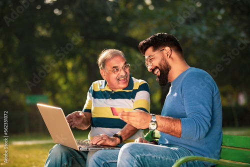 Young indian man showing bank card and laptop how to uses to his father at park