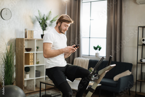Portrait of focused young male wearing sportswear using exercise bike typing message on phone. Home fitness workout sporty man training on smart stationary bike indoors.