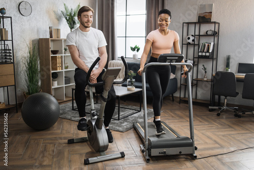 Portrait of focused caucasian male and african female wearing sportswear using exercise bike and treadmill. Home fitness workout sporty people training on exercise machines indoors.