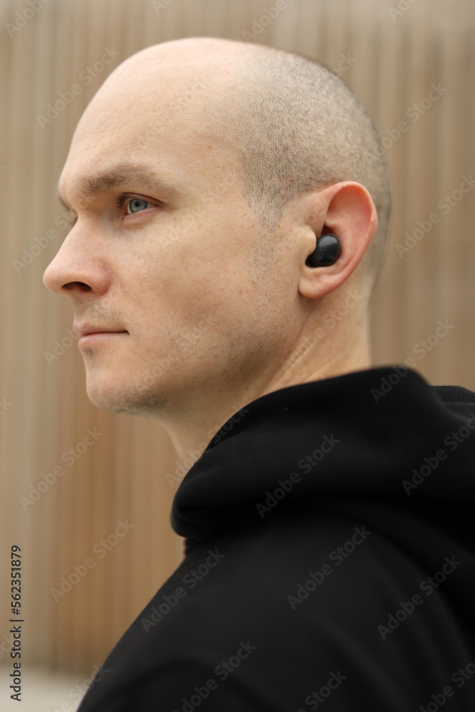 Fototapeta premium Young bald man with freckles on face. Guy wearing in black hoodie and with wireless headphones. Portrait in profile. Wooden background.