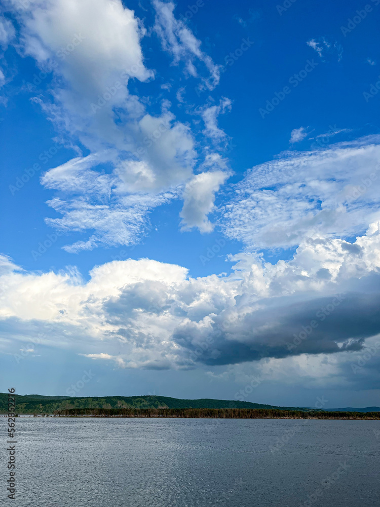 Trees and white clouds by the river with blue water. River landscape. Reflection of the sky in the river.