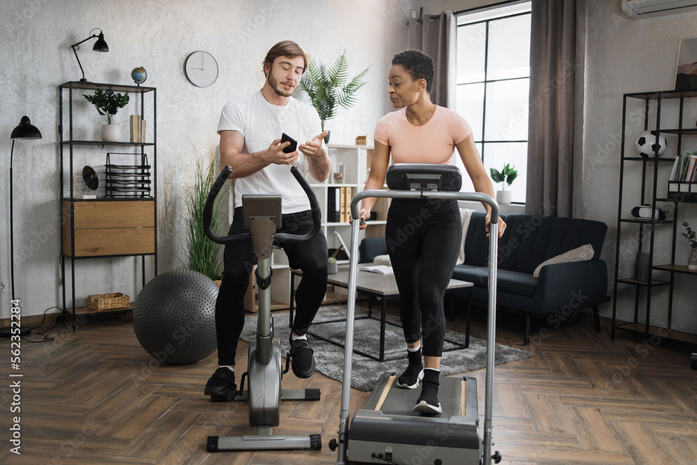 Fototapeta premium Happy smiling couple doing cardio on stationary bike and treadmill using smart phone to watch online video lessons how to sole weight. Attractive female and male working out on fitness at home.