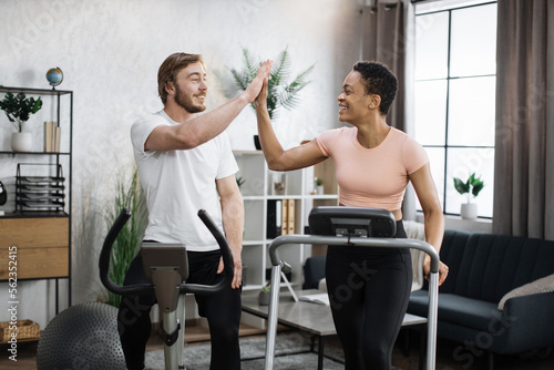 Young couple doing cardio on stationary bike and treadmill using smart phones to control physical parameters giving high five. Attractive female and male working out on fitness at home.