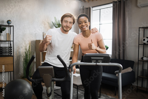 African woman running on treadmill and caucasian man training on exercise bike. Young couple doing cardio on stationary bike and treadmill. Attractive female and male working out at home.