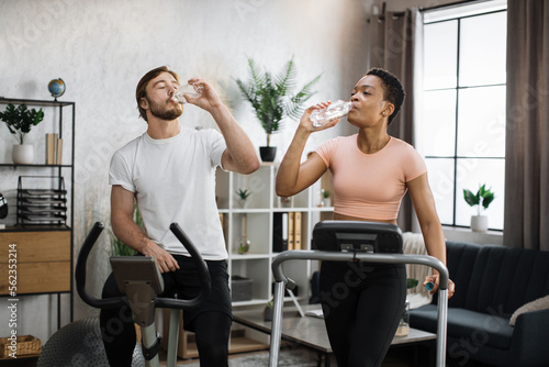 Young african american sports woman in sportswear running on treadmill and fit caucasian man cycling bike at home while drinking water from bottles  celebrating good results.