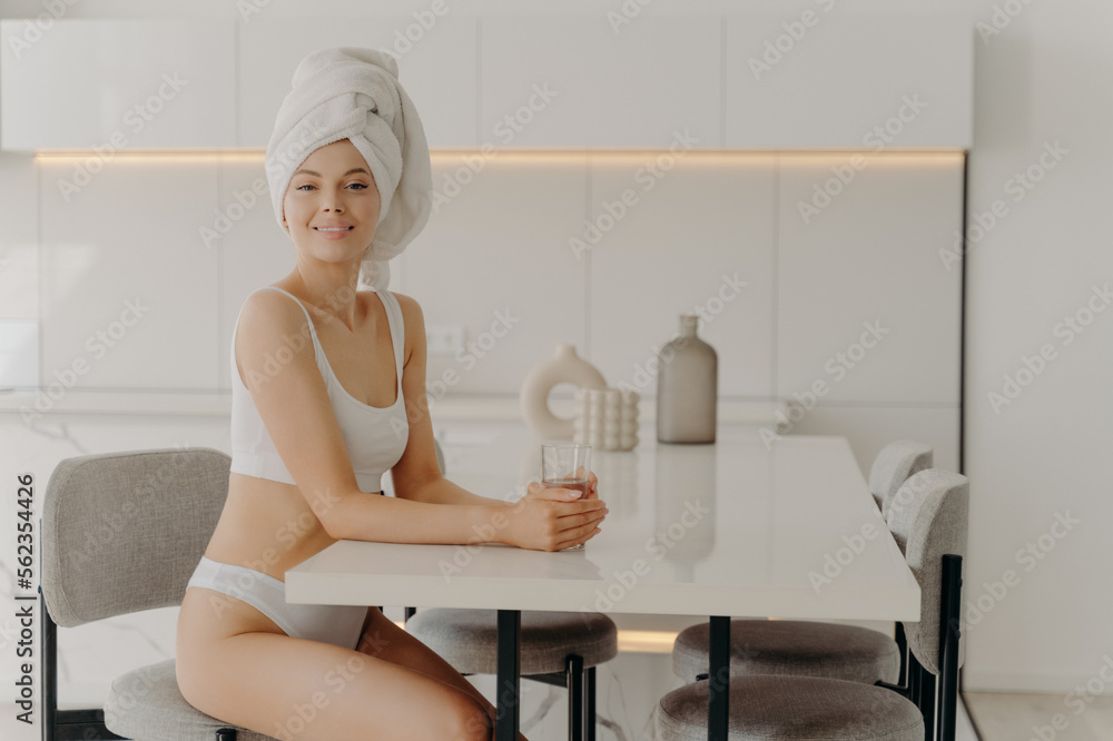 Relaxed beautiful slender woman sitting in kitchen after taking shower with glass of water Relaxed beautiful slender woman sitting in kitchen after taking shower with glass of water