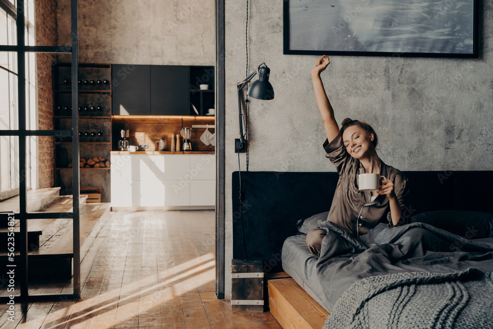 Young beautiful woman sitting on bed stretching with her hand up while holding cup of coffee Young beautiful woman sitting on bed stretching with her hand up while holding cup of coffee