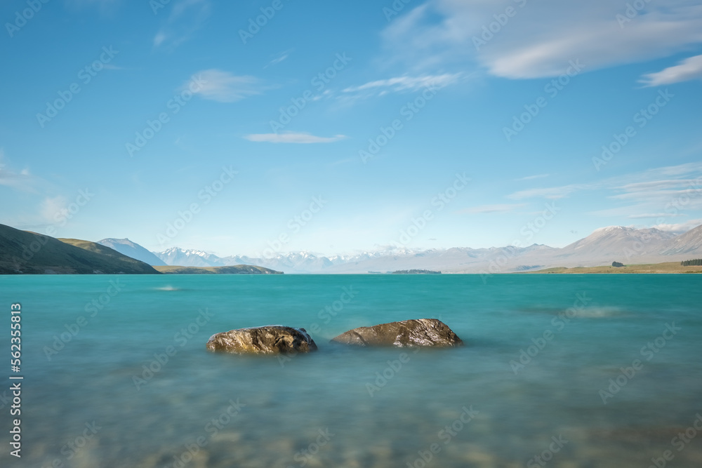 Two rocks in the turquoise blue water of Lake Tekapo in New Zealand with snow capped mountains in the distance