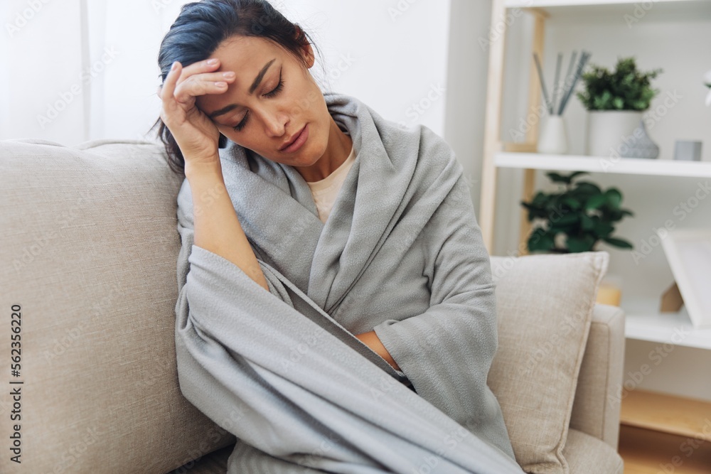 Woman cold headache with a cup of tea sits on a sofa in a plaid, treatment for flu, allergies and covid-19 Woman cold headache with a cup of tea sits on a sofa in a plaid, treatment for flu, allergies and covid-19