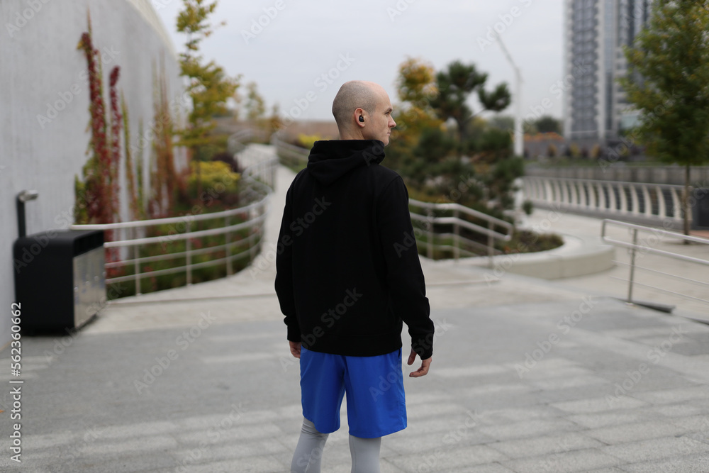 A bald male athlete in sportswear and black wireless headphones stands in a city park and looks away. Back view.
