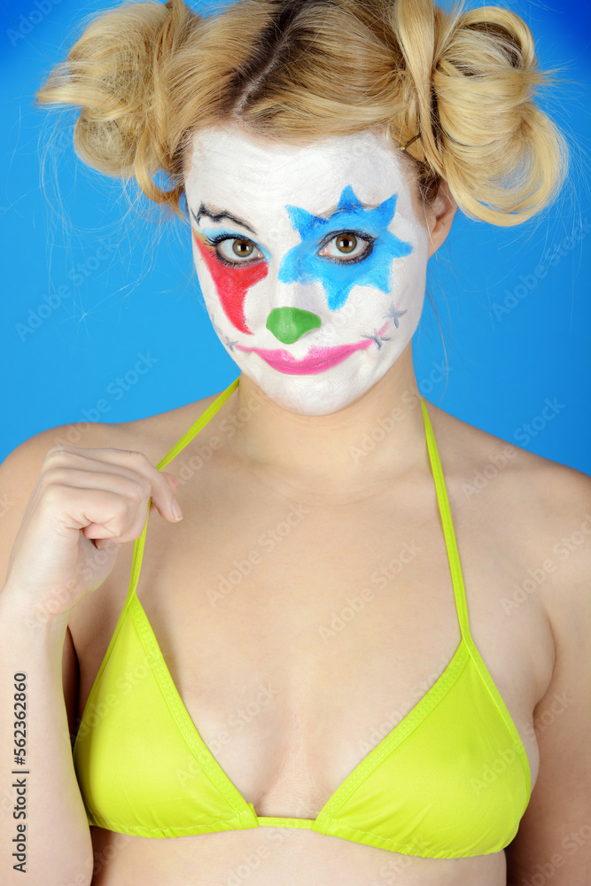 Portrait of an ugly clown in bikini grimaces and poses in front of blue background in studio