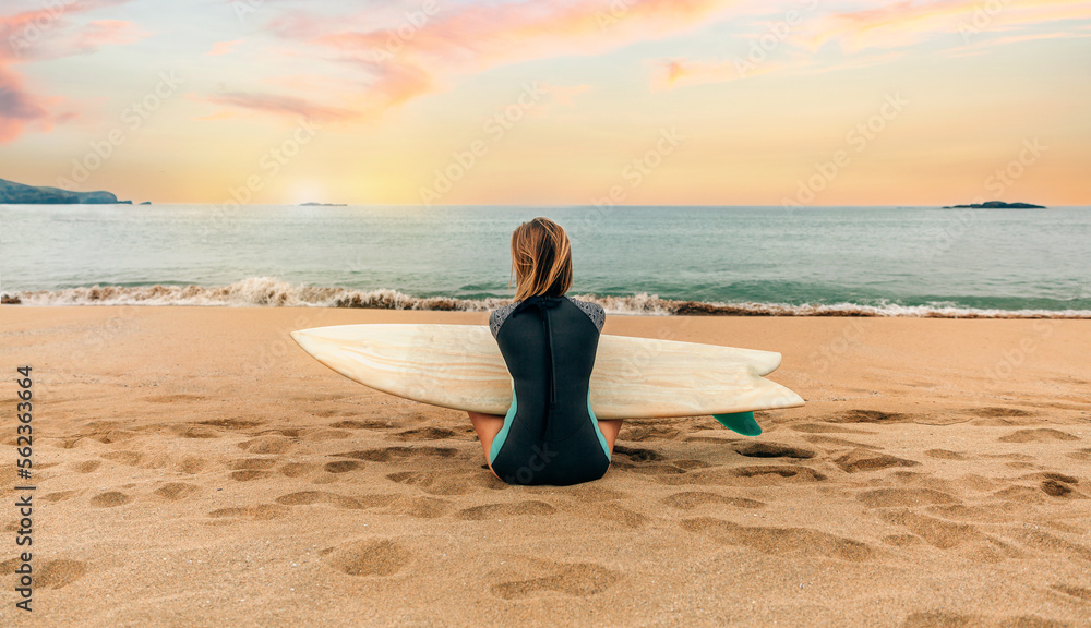 Unrecognizable young surfer woman with wetsuit and surfboard sitting on the sand looking at the sea