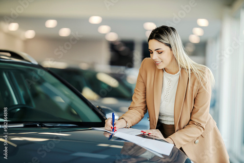 A happy customer is signing up contract and paperwork while standing at the car showroom.