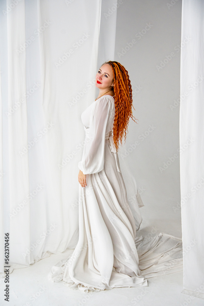 beautiful red-head girl in white long dress with long curly hair holding posing in white studio with curtains beautiful red-head girl in white long dress with long curly hair holding posing in white studio with curtains