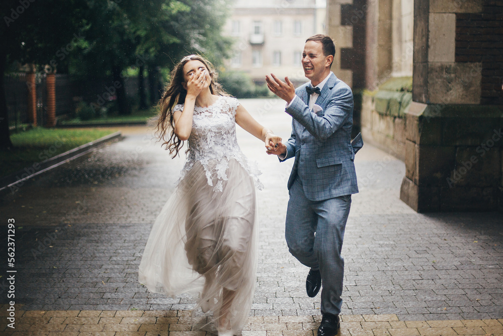 Naklejka premium Stylish happy bride and groom running on background of old church in rainy street. Provence wedding. Beautiful emotional wedding couple smiling and having fun in rain in european city.