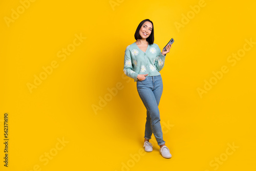 Full size photo of thoughtful girl wear blue cardigan hold phone arm in pocket look empty space isolated on yellow color background