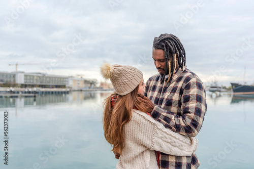 a multiracial couple standing face to face on the pier hugging each other, he touches her face