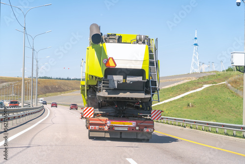 Heavy industrial truck with low side on low-frame platform transports disassembled big green combine harvester along wide highway on summer day.